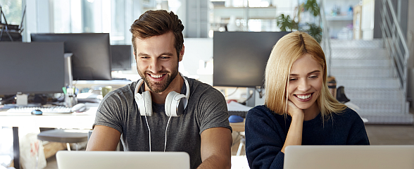 Foto: Junger Mann und junge Frau sitzen lächelnd nebeneinander vor ihren Computerbildschirmen in einem Großraumbüro. 